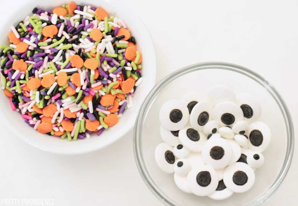 Halloween sprinkles and candy eyeballs in small ingredient bowls on a white surface.