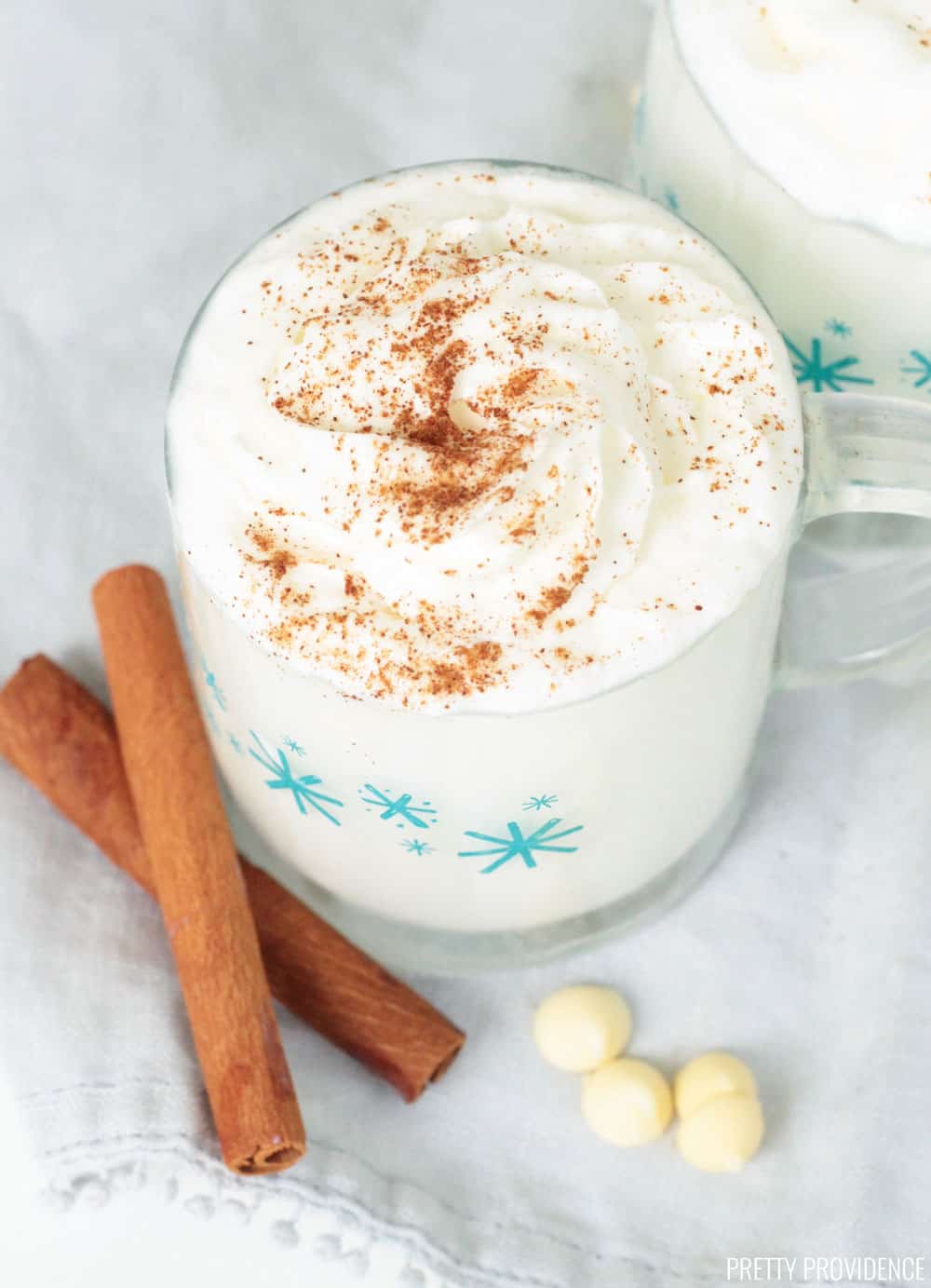 Mug of white hot chocolate topped with whipped cream and dusted with cinnamon, cinnamon sticks and white chocolate chips on the side.