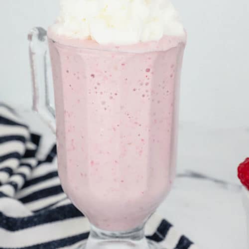 Raspberry Milkshake in a tall milkshake glass topped with whipped cream with raspberries as a garnish.