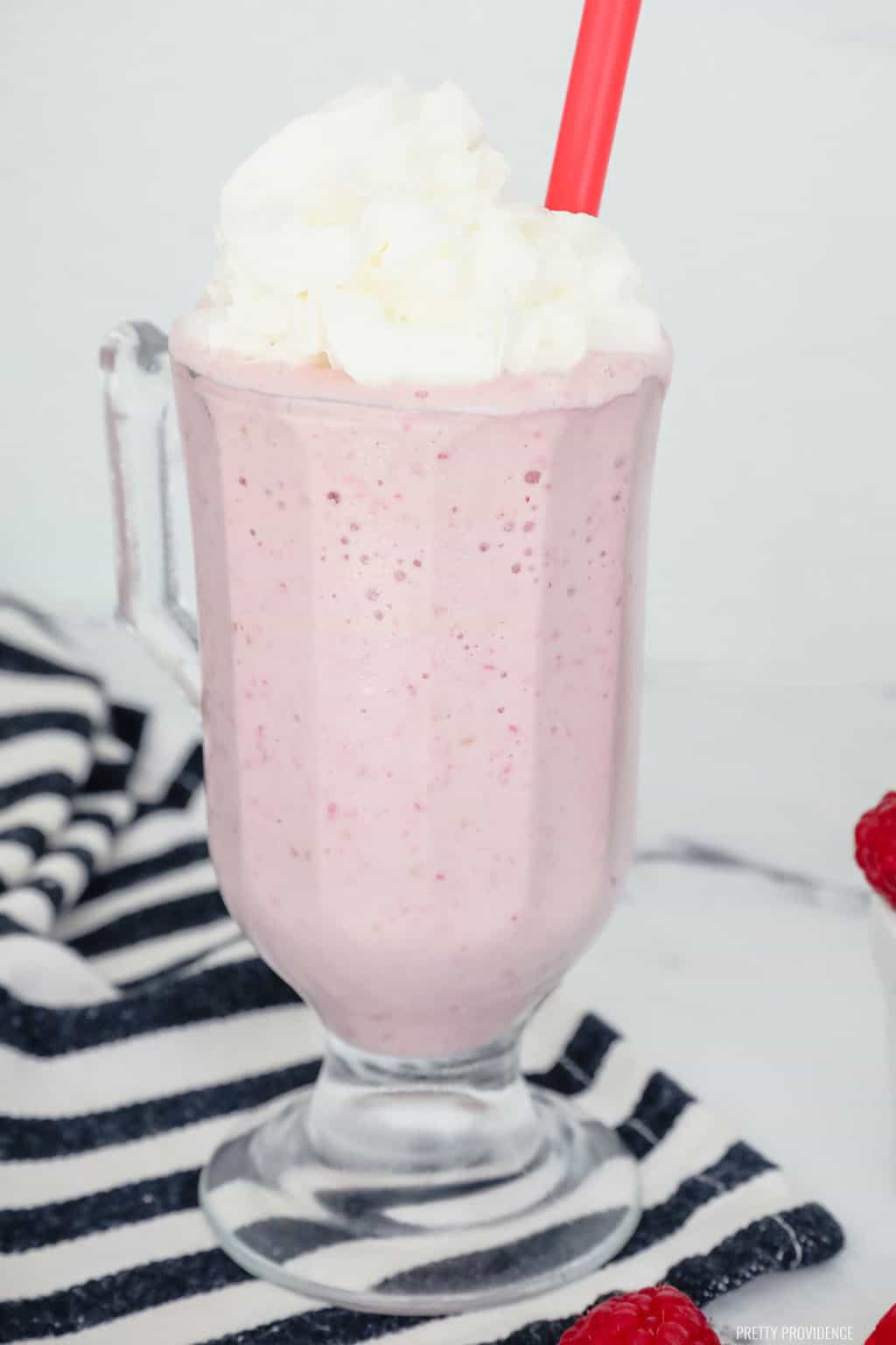 Raspberry Milkshake in a milkshake glass topped with whipped cream with fresh raspberries on the table.