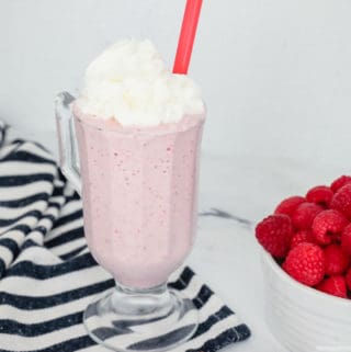 Raspberry milkshake in a milkshake glass topped with whipped cream, a bowl of fresh raspberries on the side.