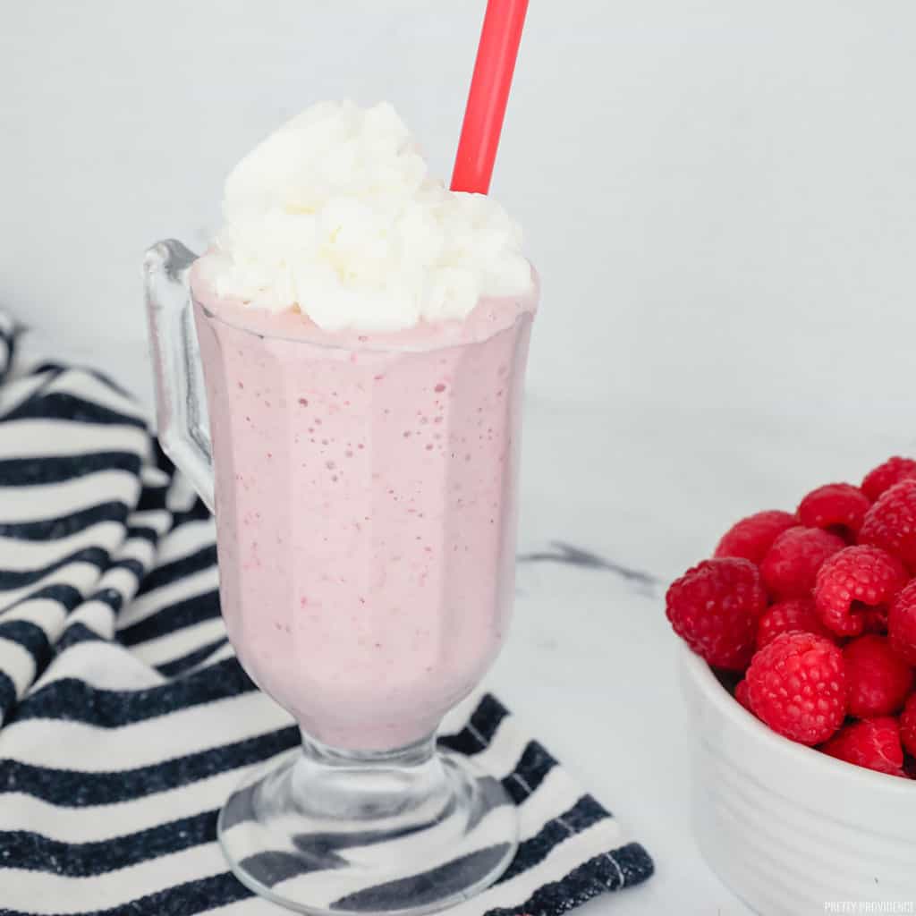 Raspberry milkshake in a milkshake glass topped with whipped cream, a bowl of fresh raspberries on the side.