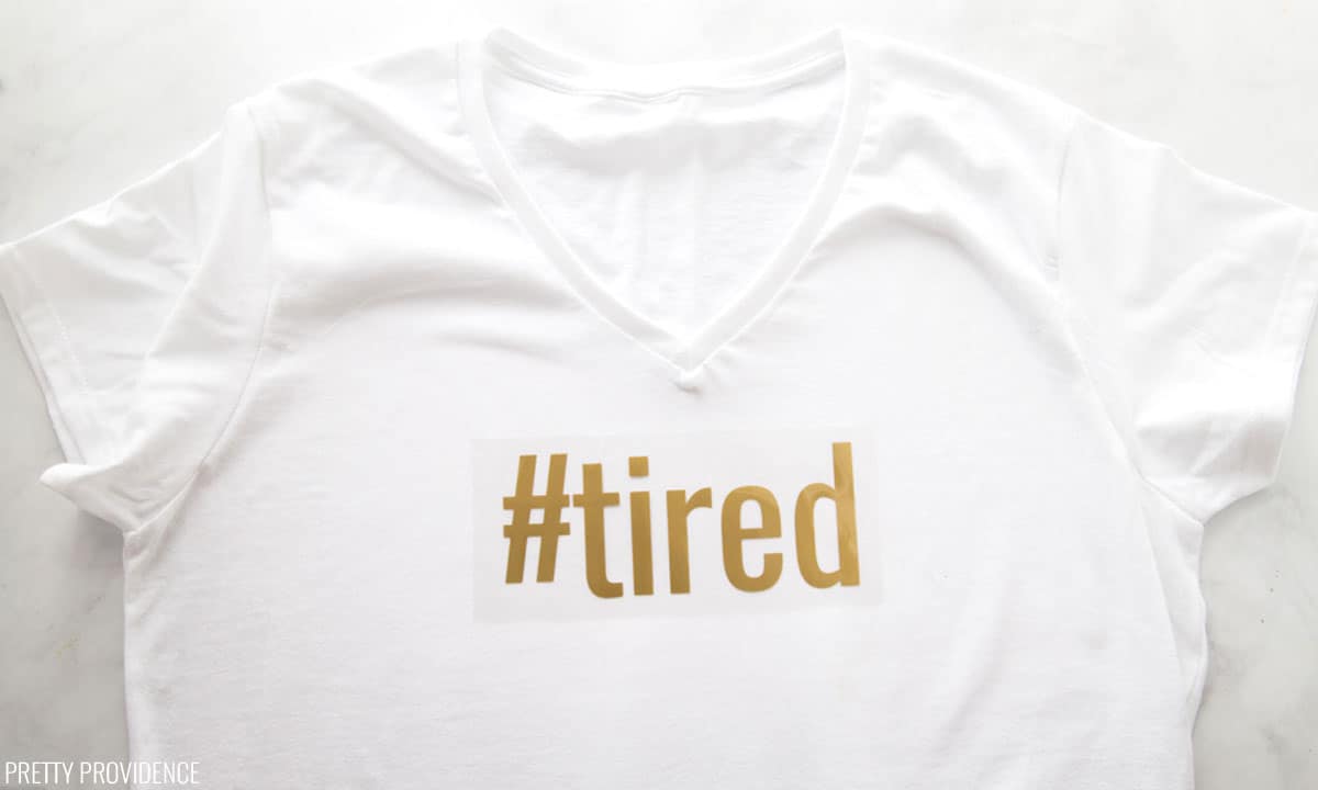 "#tired" in gold iron-on attached to plastic ready to be ironed onto a white t-shirt