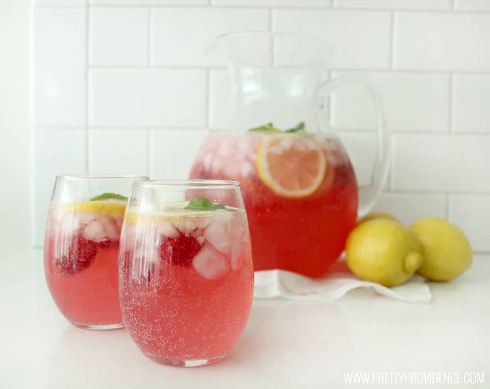 a pitcher of raspberry lemonade next to some lemons with two filled glasses of lemonade in the foreground.