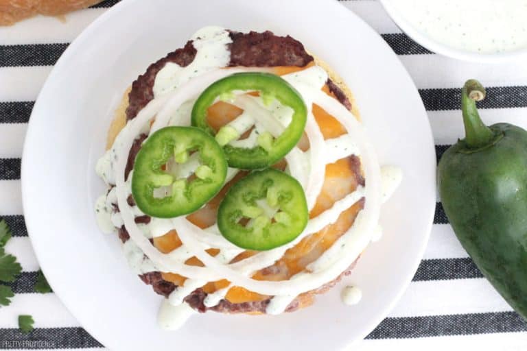 Angus beef patty on a white plate, topped with melted cheese, jalapeño ranch, onions and jalapeño peppers.