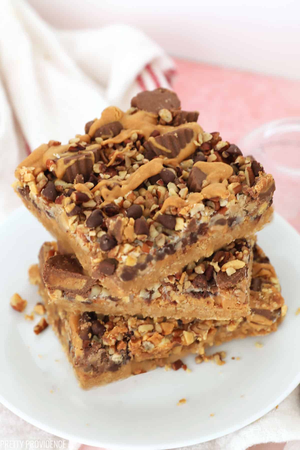 Stack of three peanut butter magic cookie bars with mini peanut butter cups, pecan and peanut pieces, mini chocolate chips and drizzled peanut butter on top.