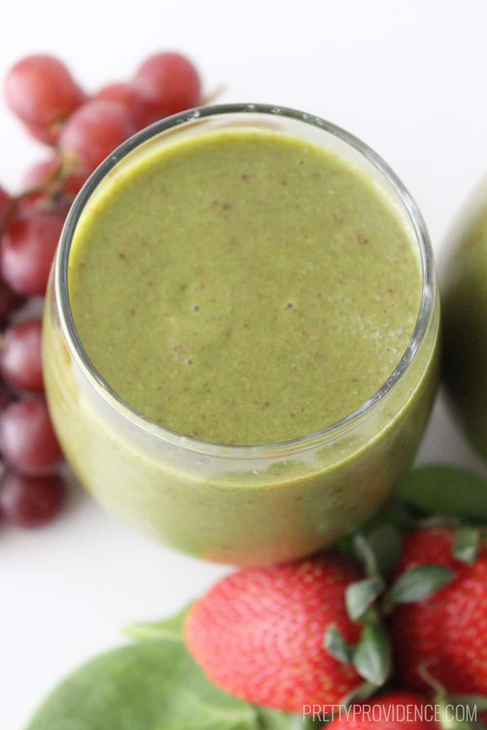 Green smoothie in a clear glass, with red grapes and strawberries on the side.