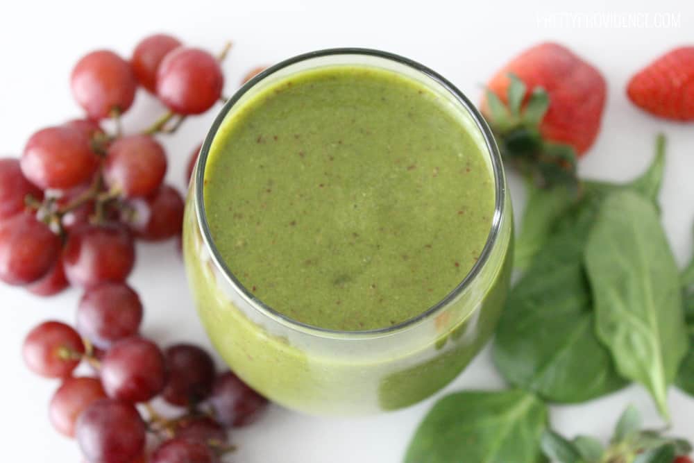 Green smoothie in a clear glass, surrounded by grapes, strawberries and spinach.