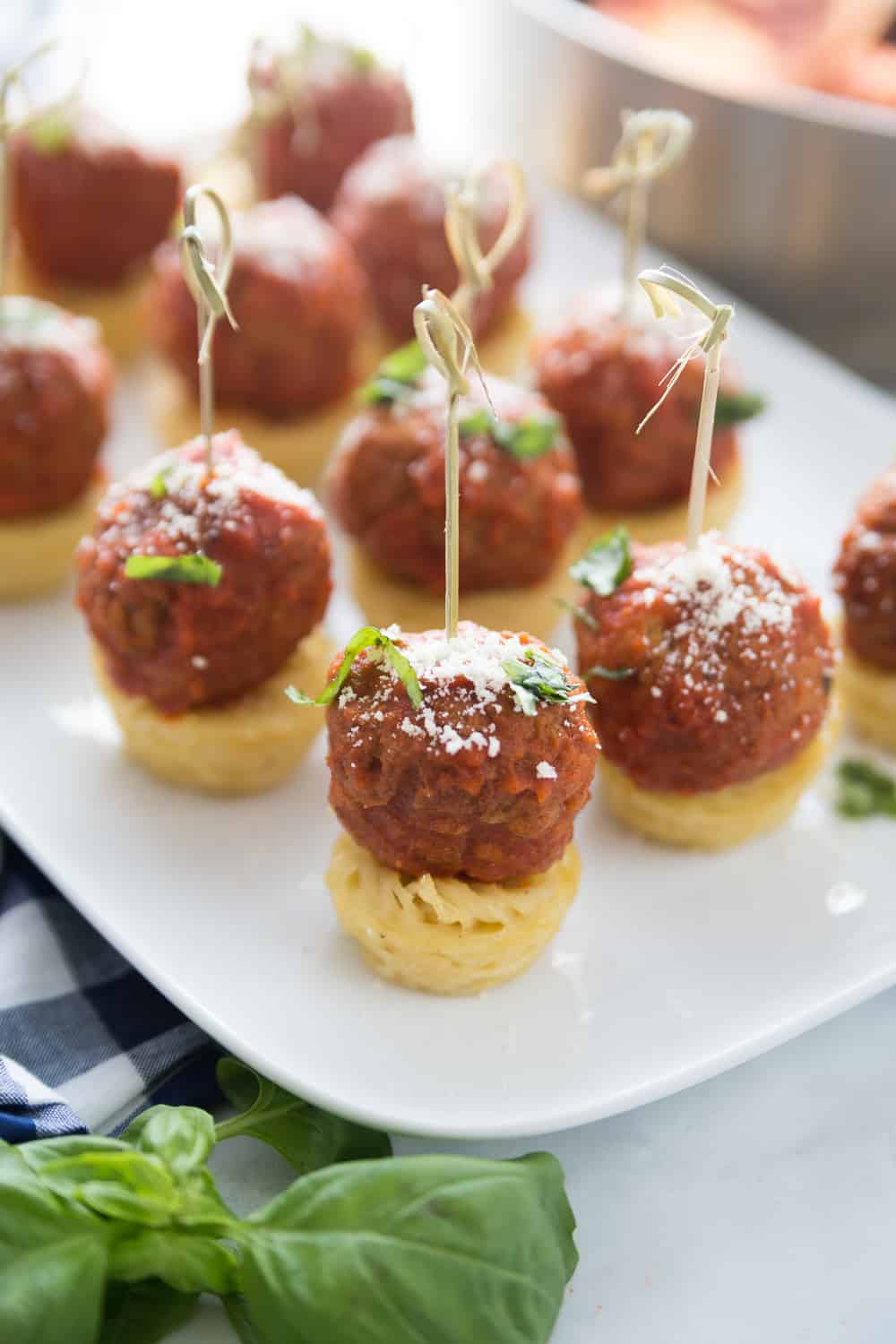Spaghetti and meatball appetizers on a white serving dish, little spaghetti muffins with meatballs on top, parmesan cheese and basil sprinkled on top and a toothpick holding them together.