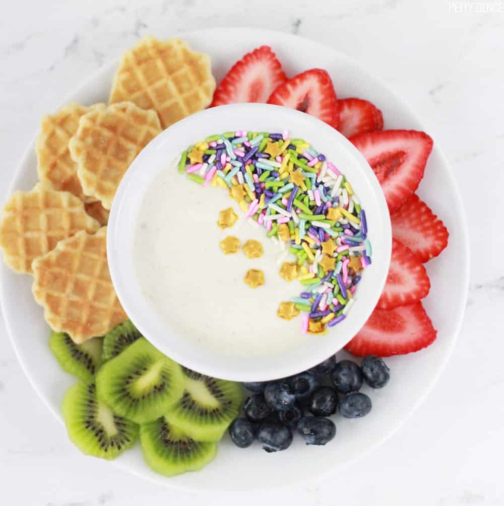 Kids yogurt with fruit and sprinkles in a white bowl, in the center of a plate of fruit with strawberries, kiwi, blueberries and waffle chips.
