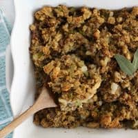 Thanksgiving stuffing in a white casserole dish with a wooden spoon full of stuffing being picked up from out of the pan.