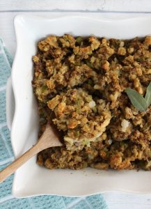 Thanksgiving stuffing in a white casserole dish with a wooden spoon full of stuffing being picked up from out of the pan.