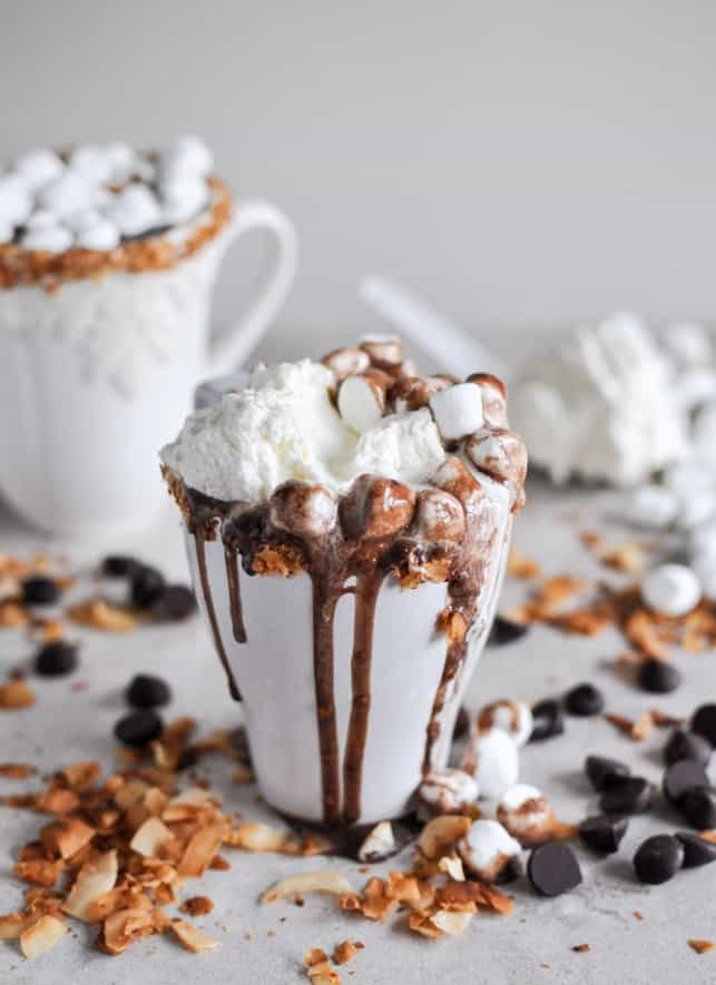 Hot Chocolate overflowing out of a white mug, topped with whipped cream, marshmallows and hot fudge sauce.
