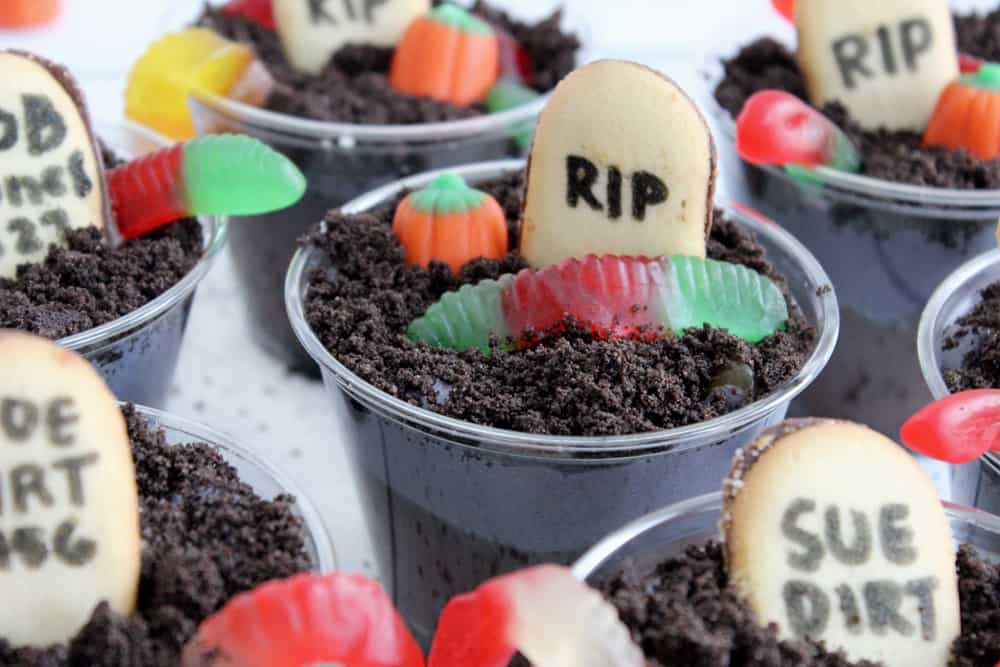 assorted dirt cups with gummy worms, pumpkins and tombstone cookies topping them
