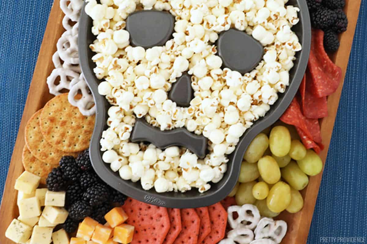 Halloween snacks like pretzels, fruit, crackers and cheese on a serving board and popcorn in a skull shaped cake pan in the middle. 