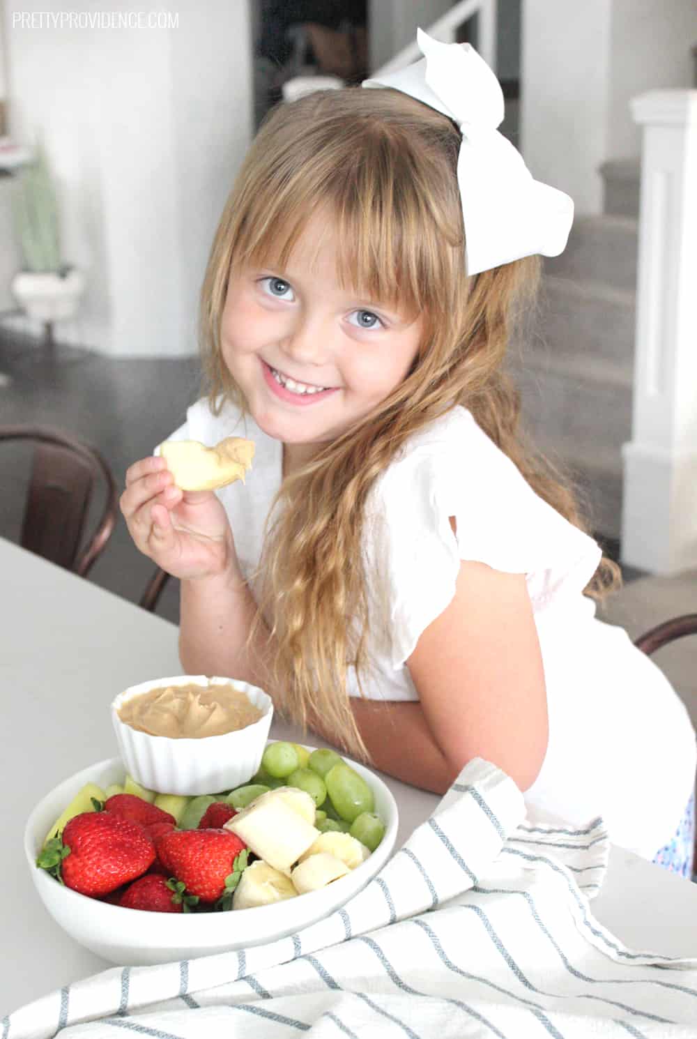 Little girl in white shirt eating strawberries, bananas, grapes and peanut butter dip.