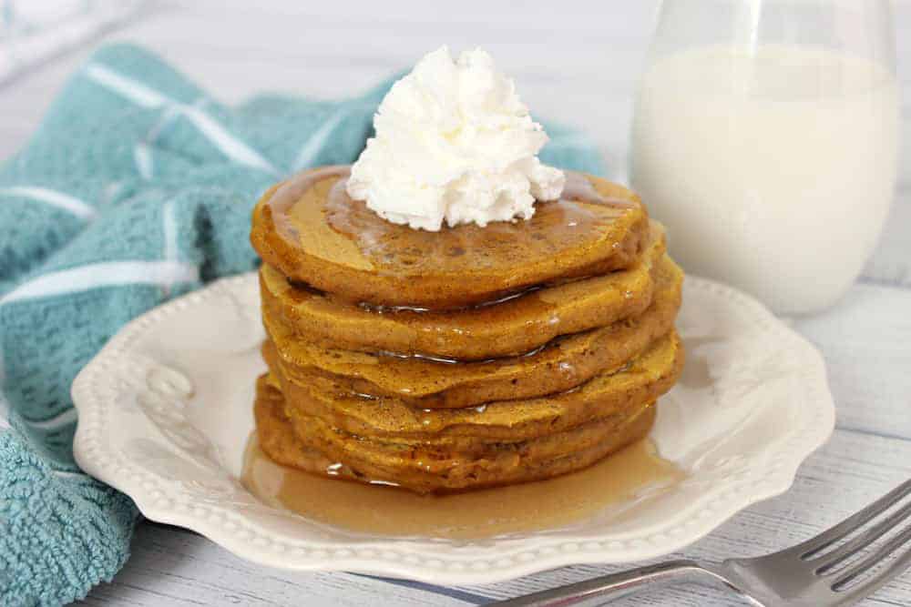 Pumpkin Pancakes stacked high on a white plate with syrup and whipped cream on top.