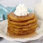 Pumpkin Pancakes stacked high on a white plate with syrup and whipped cream on top.