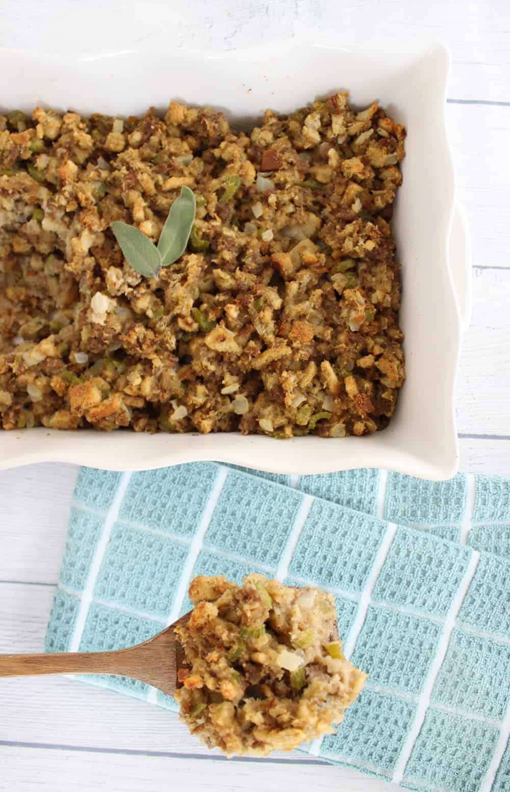 Thanksgiving Make Ahead Stuffing with sausage and herbs in a white casserole dish, a wooden spoon full of stuffing on the counter next to it.
