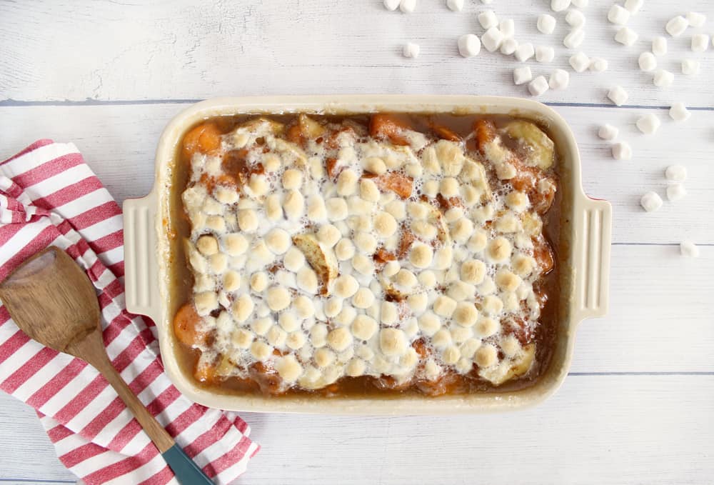 sweet potato casserole on a wood background with a red and white striped tea towel, wooden spoon, and marshmallows