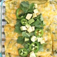 Beef enchiladas with green enchilada sauce in a glass dish topped with cilantro, jalapenos and avocado.