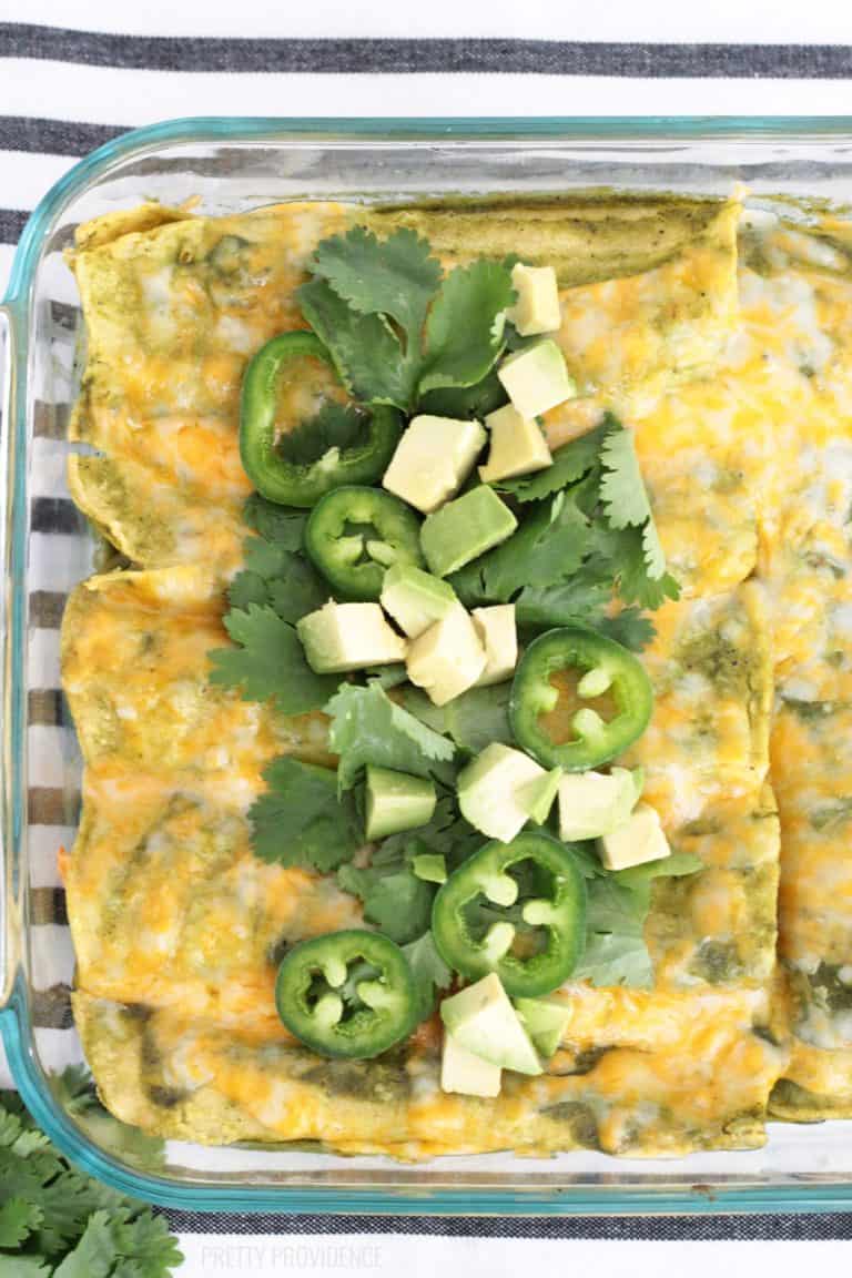 Beef enchiladas with green enchilada sauce in a glass dish topped with cilantro, jalapenos and avocado.