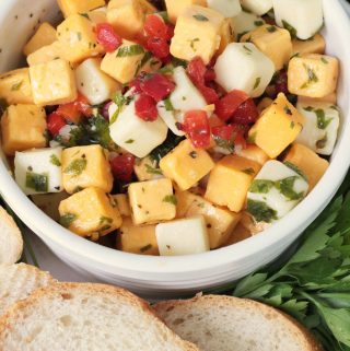 Marinated cheese in a white bowl, with yellow and white cheese cubes, pimentos, parsley, marinade and fresh baguette.