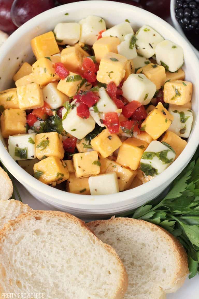 Marinated cheese in a white bowl, with yellow and white cheese cubes, pimentos, parsley, marinade and fresh baguette.