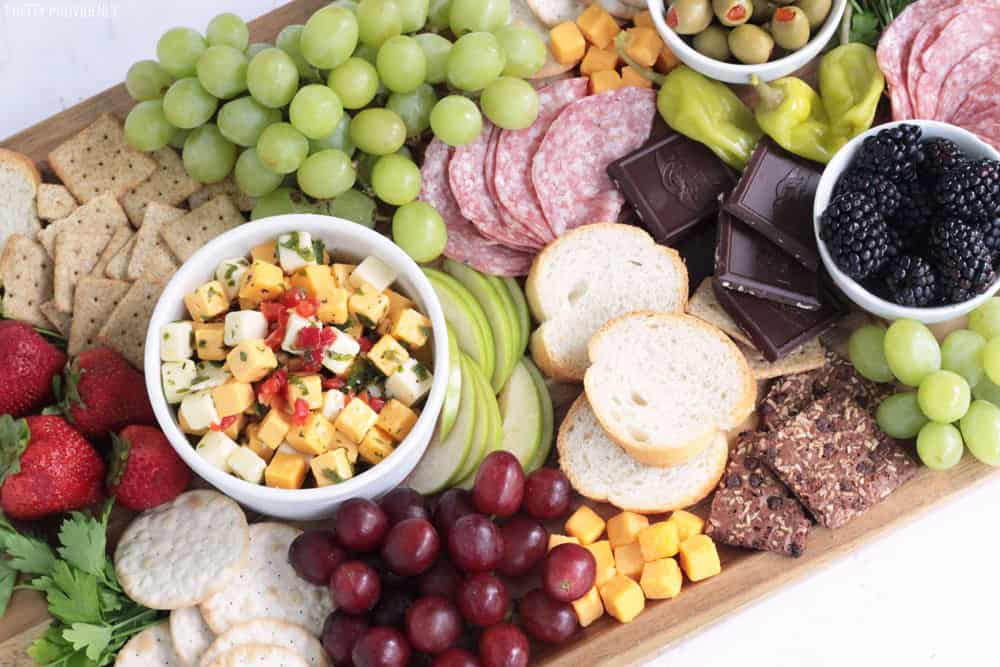 Marinated cheese in a white bowl on a cheese board with grapes, salami, bread, olives, dark chocolate, strawberries and blackberries.