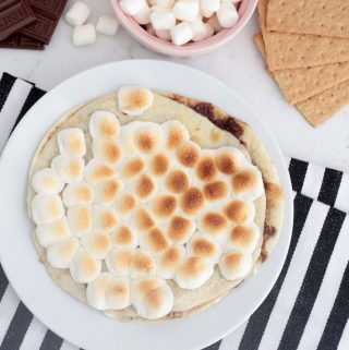 S'mores dessert quesadilla on a white plate with toasted marshmallows on top, surrounded by graham crackers, hershey bar and graham crackers.