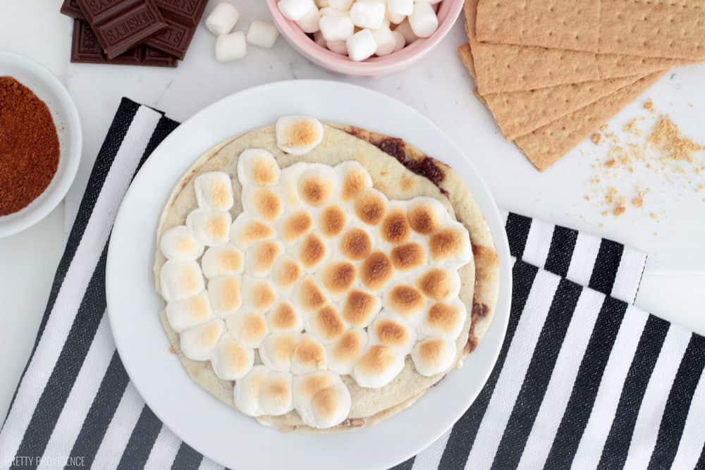 S'mores dessert quesadilla on a white plate with toasted marshmallows on top, surrounded by graham crackers, hershey bar and graham crackers.
