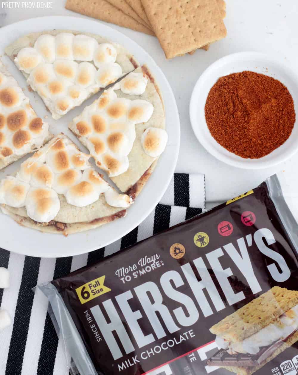 S'mores Quesadilla with toasted marshmallows on a white plate, over a striped towel, with Hershey's bars, cayenne pepper and graham crackers surrounding it.