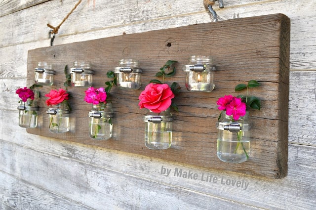 Wood plank hanging with repurposed baby food jars attached being used as flower vases and candle holders, on an outdoor wall.