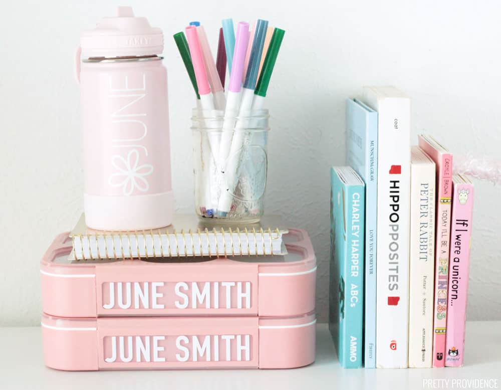 Pink bento lunch boxes, a gold notebook, pink water bottle labeled 'JUNE' crayola markers, and pastel colored board books on a white table. 