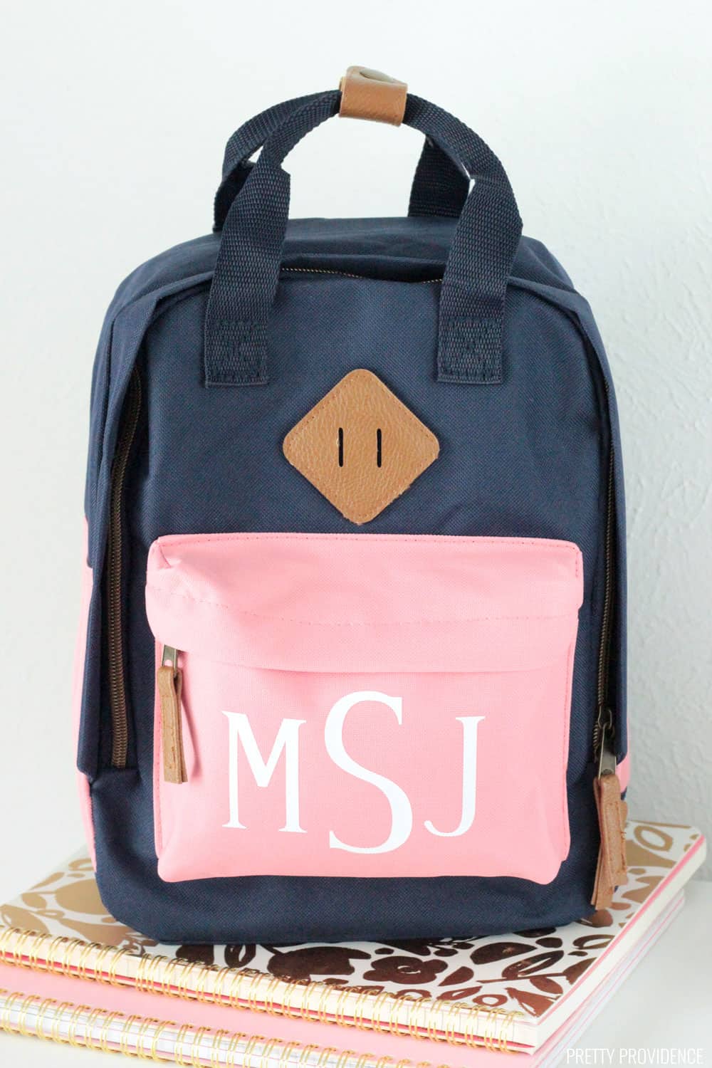 Navy blue backpack with a pink pocket monogrammed with letters M S J sitting atop two notebooks.