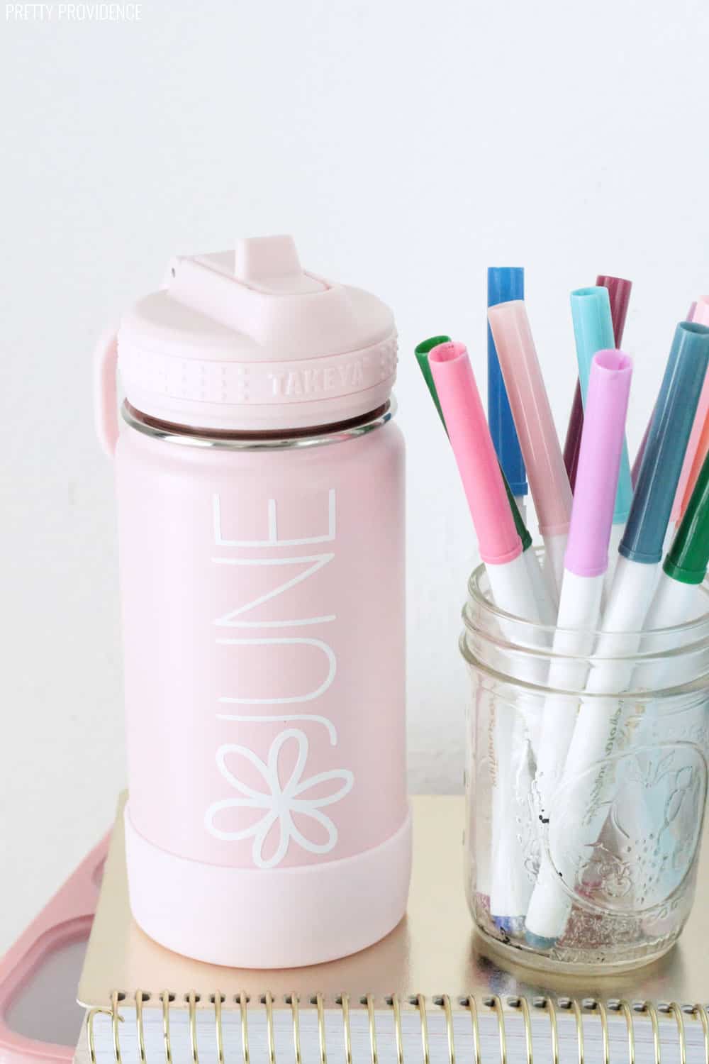 Pink water bottle labeled 'JUNE' with a flower on it, next to a jar of crayola markers, both sitting on top of a gold spiral notebook.