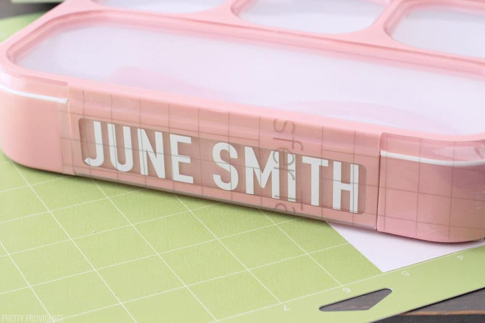 Pink bento lunch box with a white label that reads 'June Smith' and Cricut transfer tape over it, all sitting on a green StandardGrip Mat.