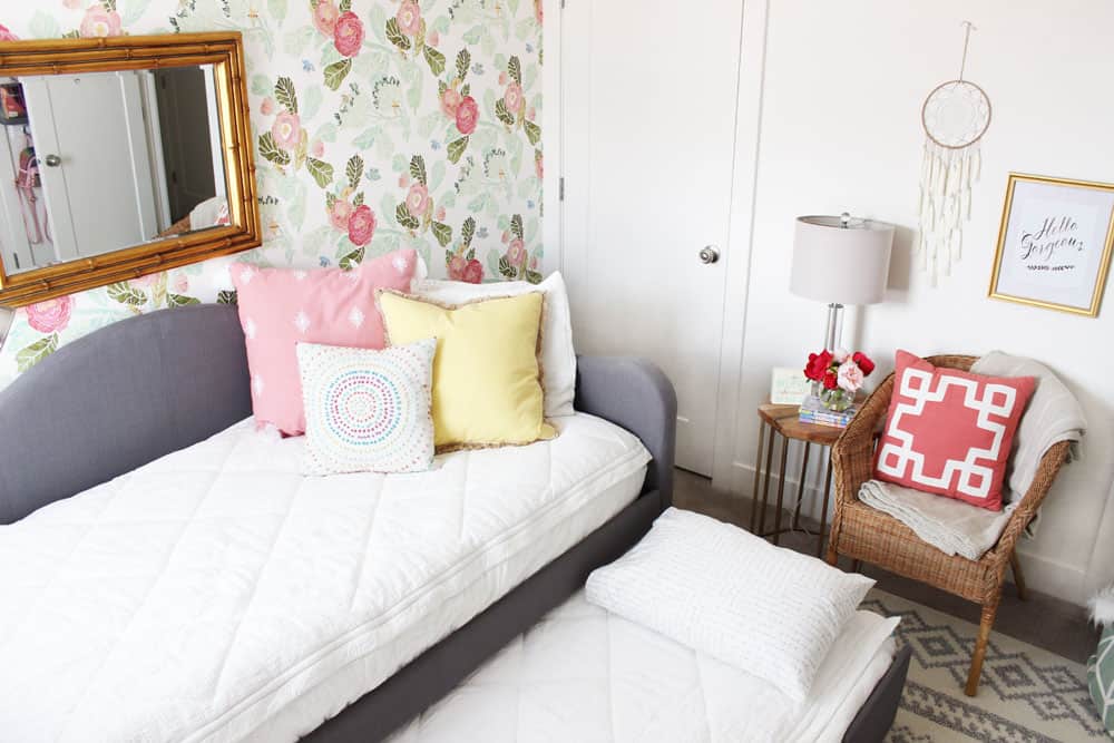 grey trundle with white bedding, a wicker chair and floral wallpaper