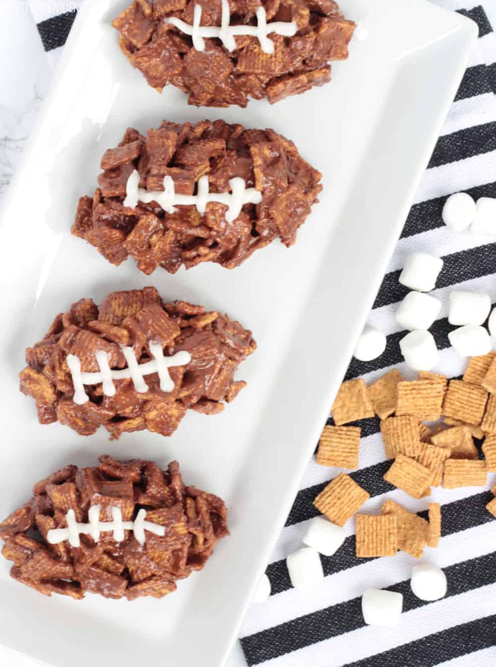Three football-shaped desserts on a white plate, s'mores bars made with golden grahams cereal, chocolate and icing.