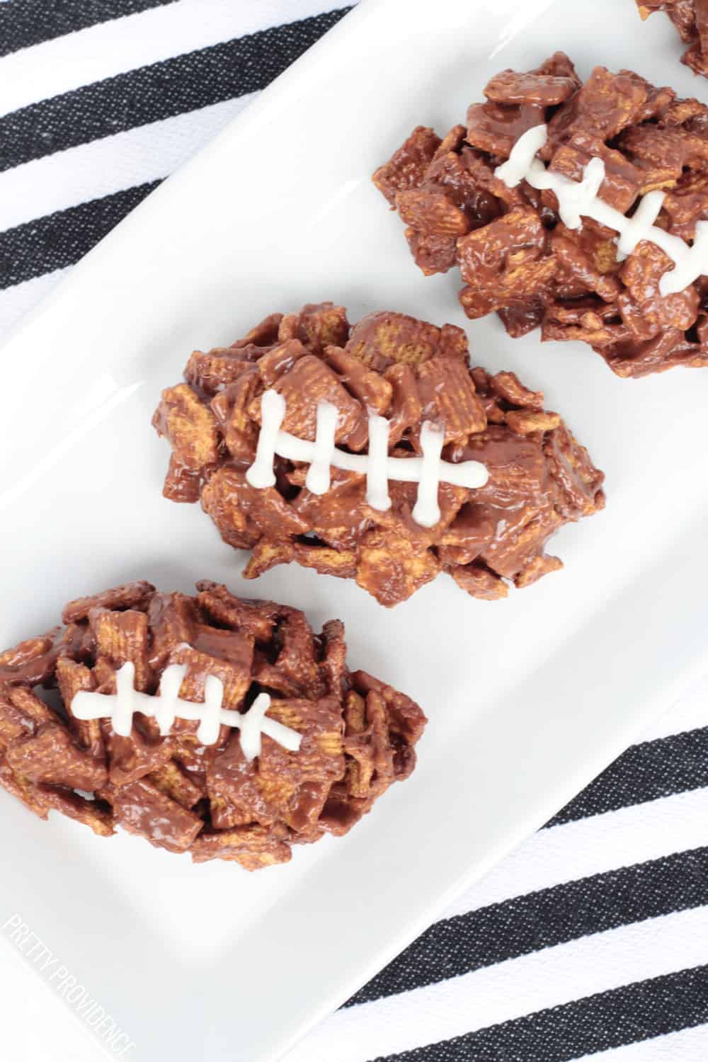 Three football-shaped desserts on a white plate, s'mores bars made with golden grahams cereal, chocolate and icing.