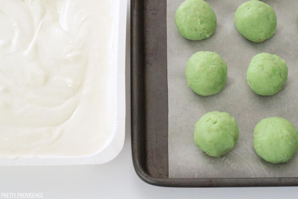 Prepared green cake balls on a cookie sheet and parchment paper next to a bowl of white melting candy.