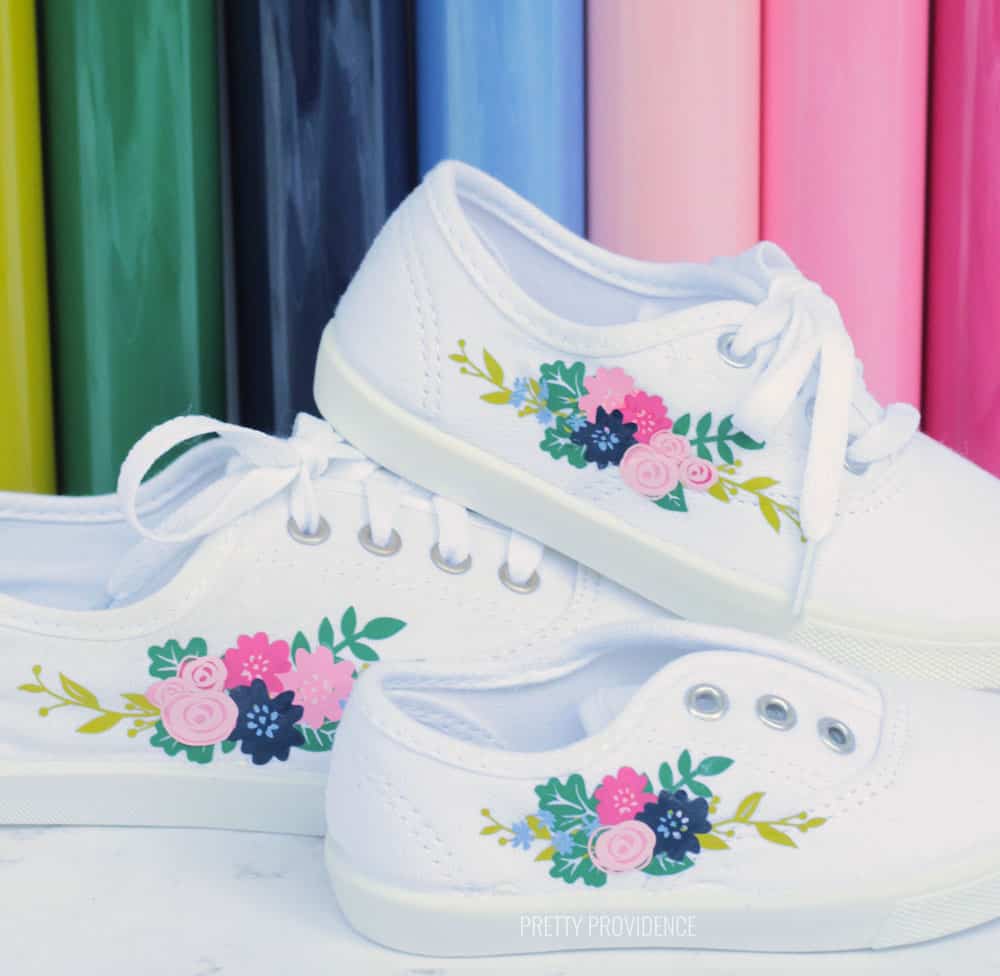 Three white shoes with floral motifs on the side, with rolls of colorful Iron On vinyl in the background. 