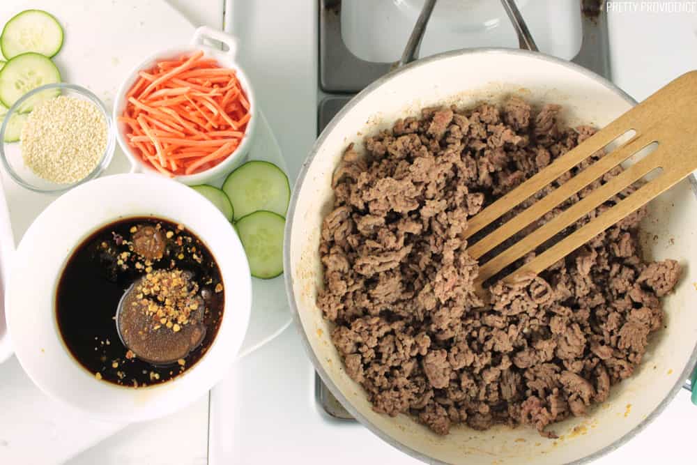 Browned lean ground beef in a pan on the stove, with Korean sauce and veggies on the side.