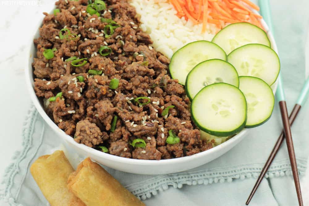 Korean beef and rice in a bowl with cucumbers, carrots on top, egg rolls on the side and chop sticks to eat with.