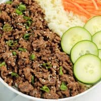 Korean Beef and rice in a bowl with green onions and sesame seeds on top, cucumbers and shredded carrots on the side.