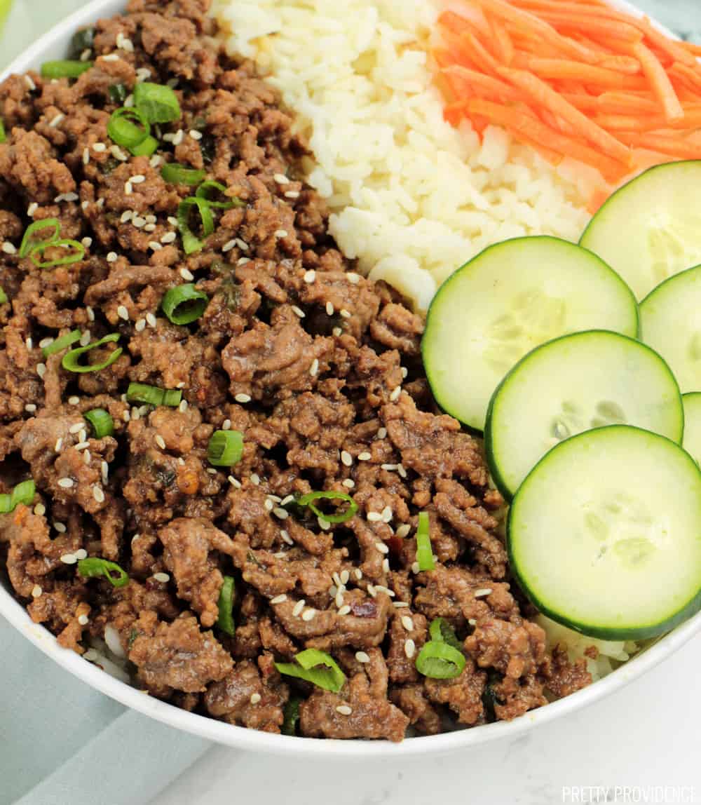 Korean Beef and rice in a bowl with green onions and sesame seeds on top, cucumbers and shredded carrots on the side.