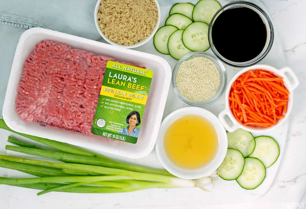 Lean Ground Beef in package, green onions, sesame oil, soy sauce, carrots and cucumbers on a marble countertop.