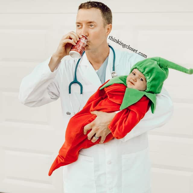 Man wearing a white doctor coat, stethoscope, and holding a baby in a pepper costume.