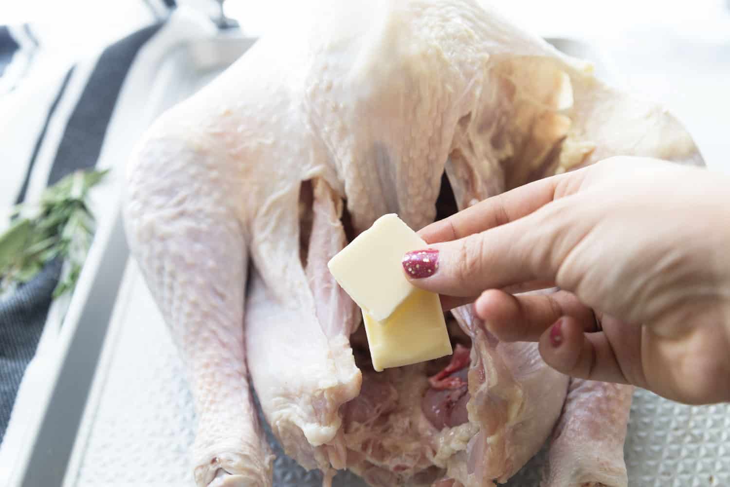 an uncooked thanksgiving turkey and a hand holding two pats of butter, preparing to put them under skin of turkey