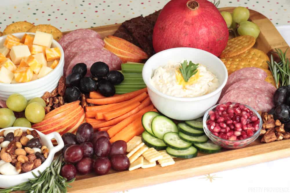 Appetizer board with vegetables, apple slices, grapes, pomegranate seeds, veggie dip, crackers and cheese cubes.